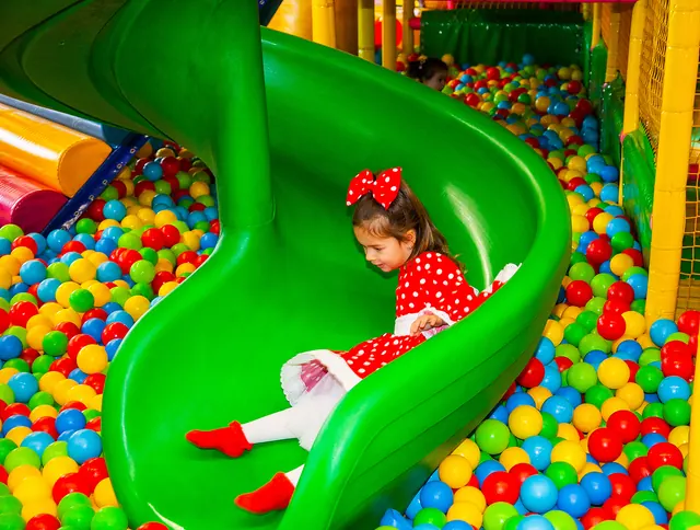 Un copil coboară pe un tobogan verde în spirală într-o piscină cu bile multicolore.